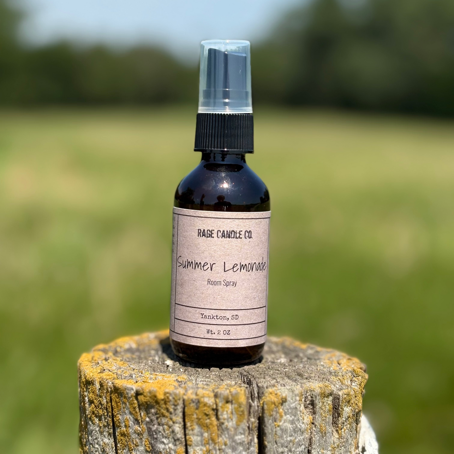 Bottle of 'Summer Lemonade' room spray on a wooden post with a blurred green field background